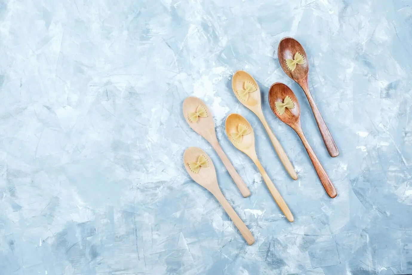 Draufsicht auf Farfalle-Nudeln in Holzlöffeln auf verputztem Untergrund, horizontal.